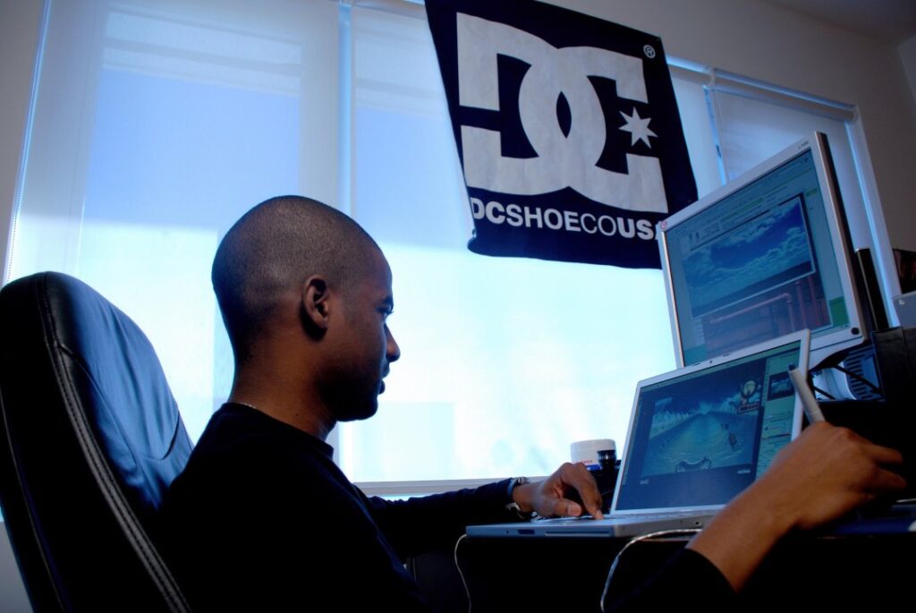 A person sits at a desk using a stylus on a tablet, with two computer screens displaying digital graphics. A DC Shoes flag hangs in the background as bright natural light enters through the window, enhancing the interactive digital experience.
