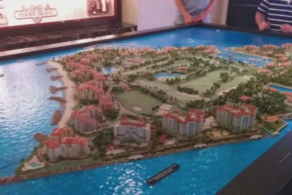 A large, detailed miniature model of a coastal resort area with buildings, green spaces, waterways, and surrounding shores showcases luxury real estate branding. Three people stand around the model.