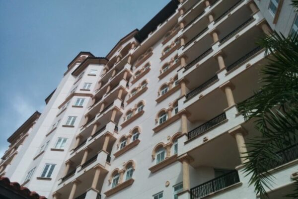 The image shows the exterior of a tall, multi-story residential building with balconies on each floor, viewed from a low angle against a partly cloudy sky, epitomizing luxury real estate branding.