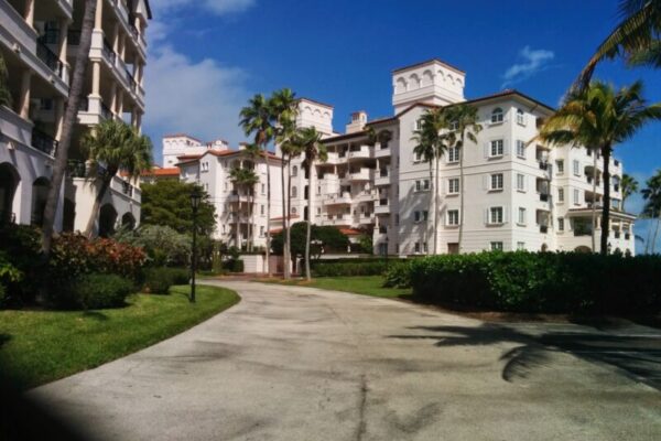 A paved path runs through a landscaped area with palm trees, leading to a large, multi-story white building complex with balconies, epitomizing luxury real estate. The clear blue sky enhances the upscale vibe of this exclusive property.