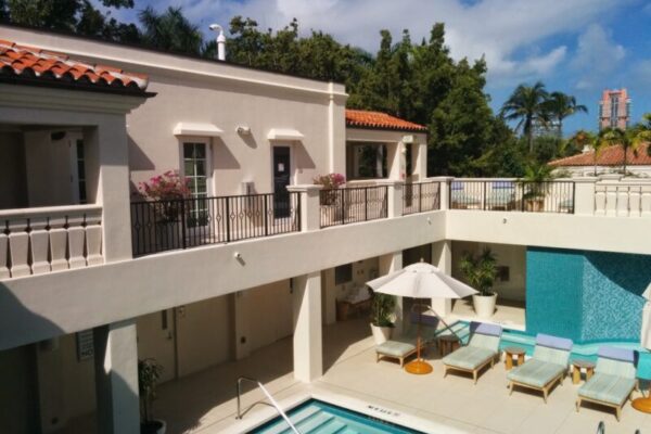 Discover the epitome of luxury real estate with this outdoor pool area featuring lounge chairs, an umbrella, and a stunning two-story white building. The upper level boasts balconies adorned with potted plants, all surrounded by lush greenery and a clear blue sky.