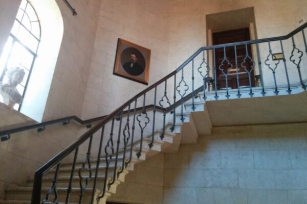 A stone staircase with a decorative wrought-iron railing leads to a wooden door, capturing the essence of luxury real estate branding. A large arched window and a framed portrait adorn the wall, while a statue bust graces the window ledge.