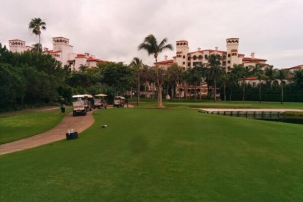 Golf course with a pathway, golf carts, and maintenance equipment, lush greenery, and palm trees. In the background, a large residential or hotel building exemplifies luxury real estate branding. The sky is overcast.