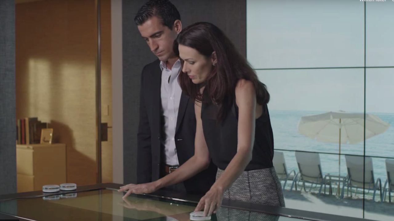 Two individuals stand by a touchscreen table in a modern office. A beach scene is visible through large windows in the background. They appear focused on the screen in front of them.