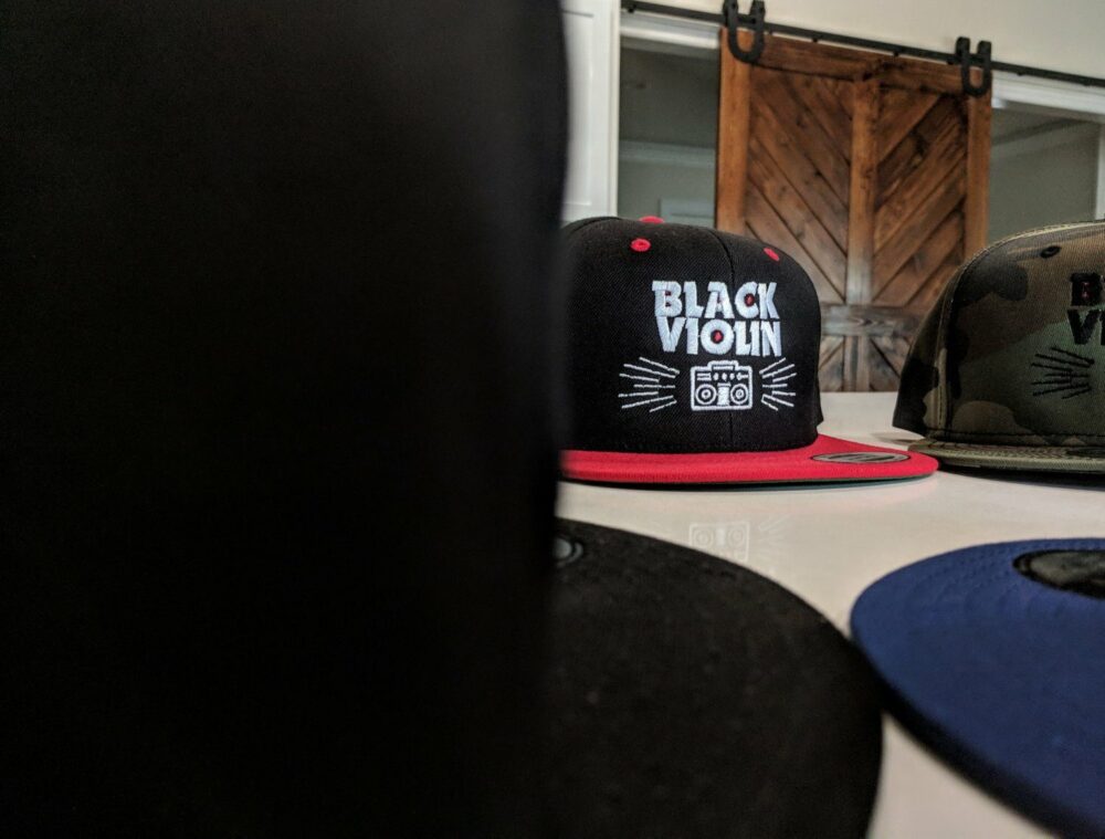 Black Violin branded baseball caps are displayed on a table, with a black violin leaning against a rustic wooden door in the background.