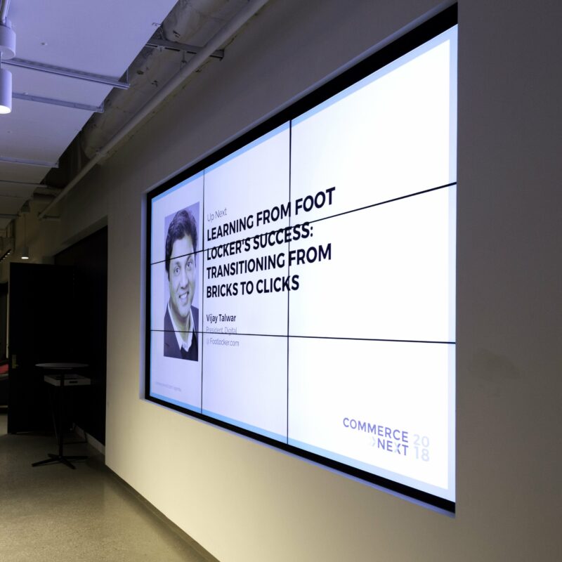 A digital screen displays a presentation titled "Learning From Foot Locker's Success: Transitioning From Bricks to Clicks" by Vijay Talwar at Commerce Next 2018.