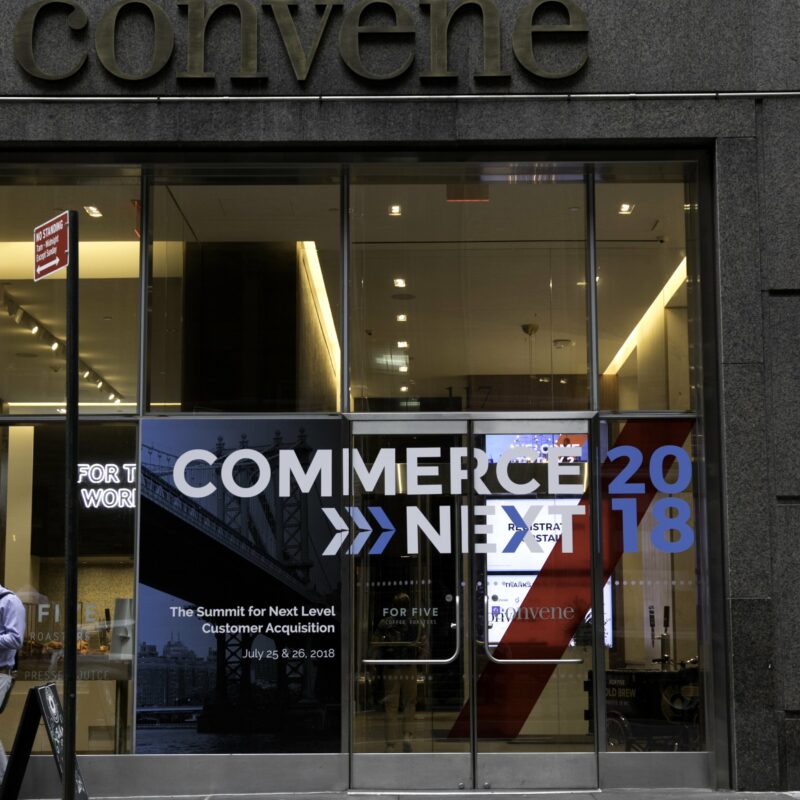An entrance of a building displaying a banner with "Commerce Next 2018," advertising a summit on customer acquisition held on July 25-26, 2018. The word "Convene" is partially visible at the top.