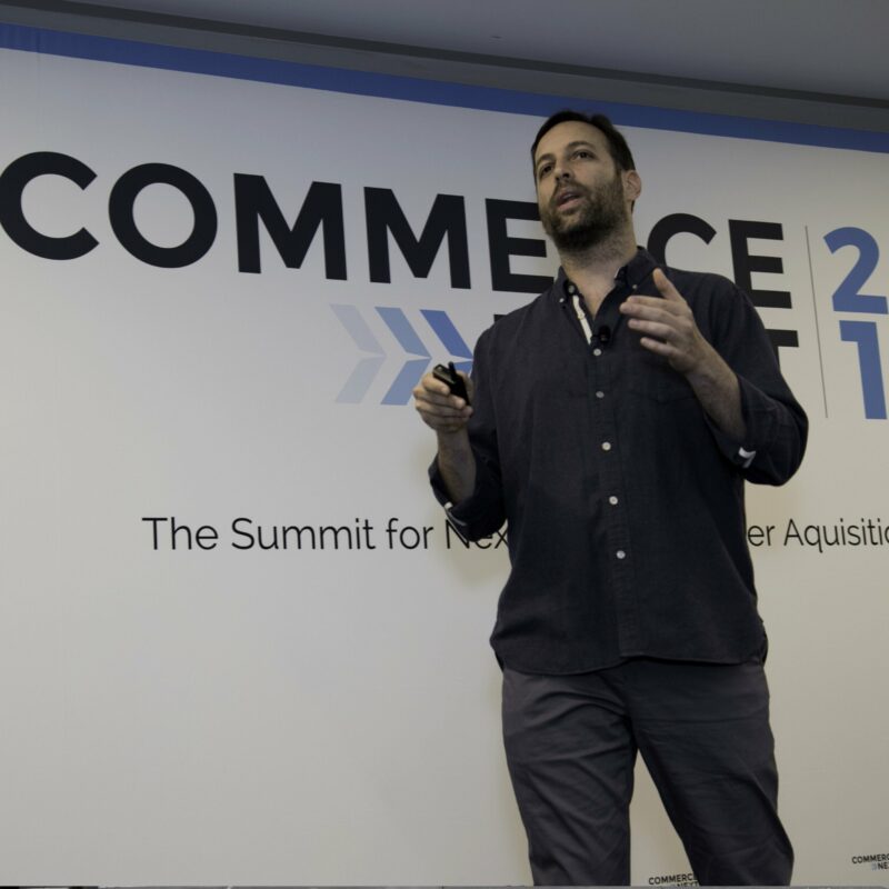 A man standing and speaking in front of a presentation backdrop that reads "COMMERCE 2021: The Summit for Next-Gen Customer Acquisition." He is holding a pointer.