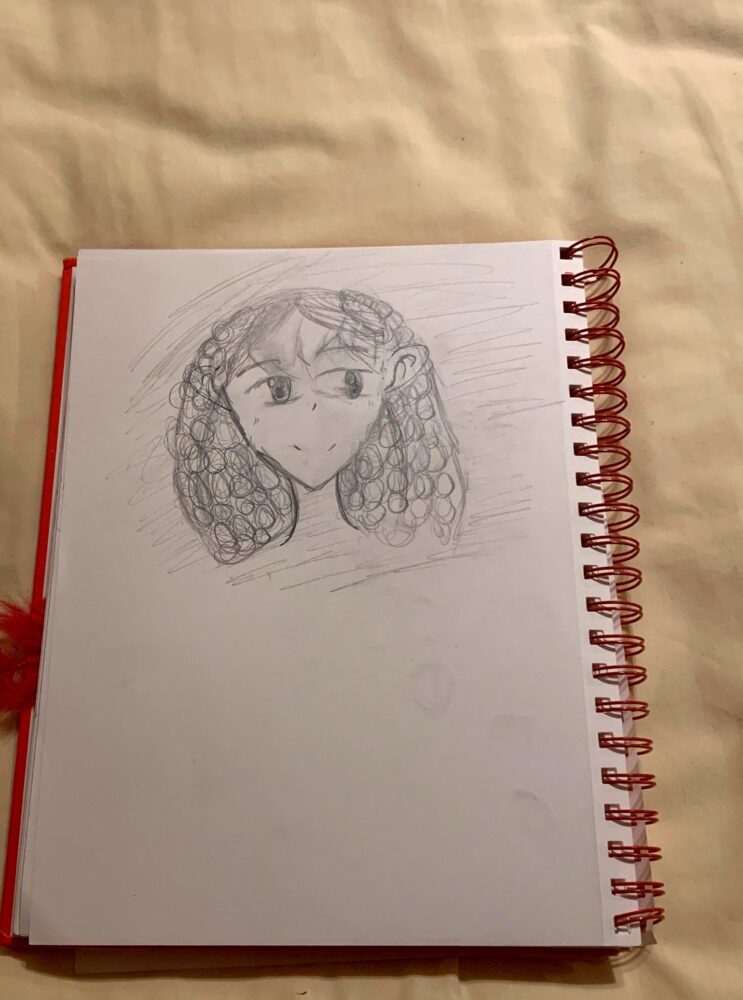 A sketch of a girl with curly hair and large eyes, drawn meticulously with a pencil on a piece of spiral-bound paper, lies gracefully on a beige surface.