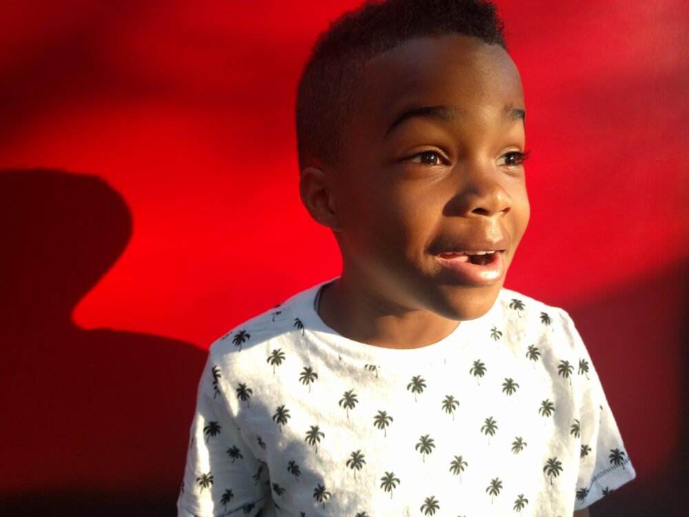 A child with a short haircut is smiling and looking off-camera, holding Encil the Pencil while wearing a white shirt with black palm tree prints. The background is a vibrant red.