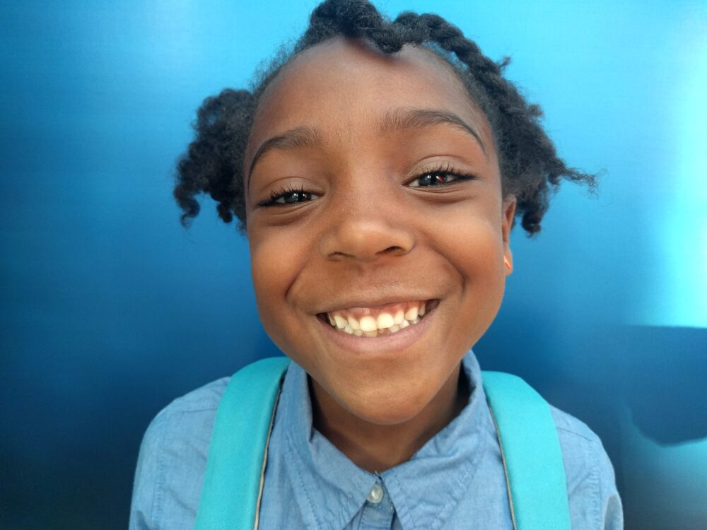 A child with braided hair and a wide smile looks directly at the camera, wearing a blue button-up shirt and a backpack, holding Encill the pencil against a blue background.