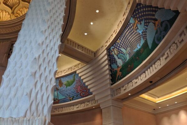 Interior view of a building with a decorative column and mosaic art on the walls, depicting various colorful scenes under an ornate ceiling.