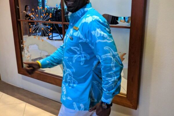 A person in a blue patterned shirt and white pants smiling and looking back, walking indoors with a decorative glass wall in the background.