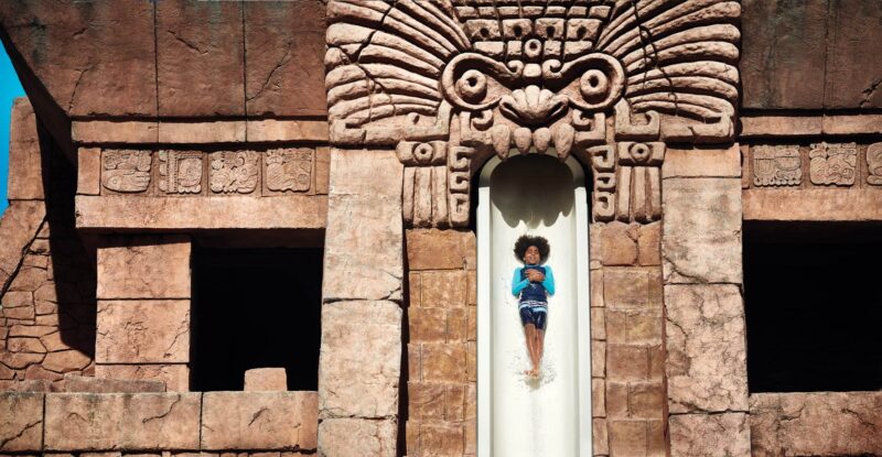 Child in blue swimwear sliding down a water slide shaped like an ancient stone temple with carved animal face at an amusement park.