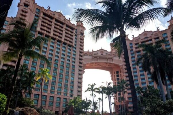 Tall, pink resort buildings connected by an archway with palm trees in the foreground and a partly cloudy sky in the background.