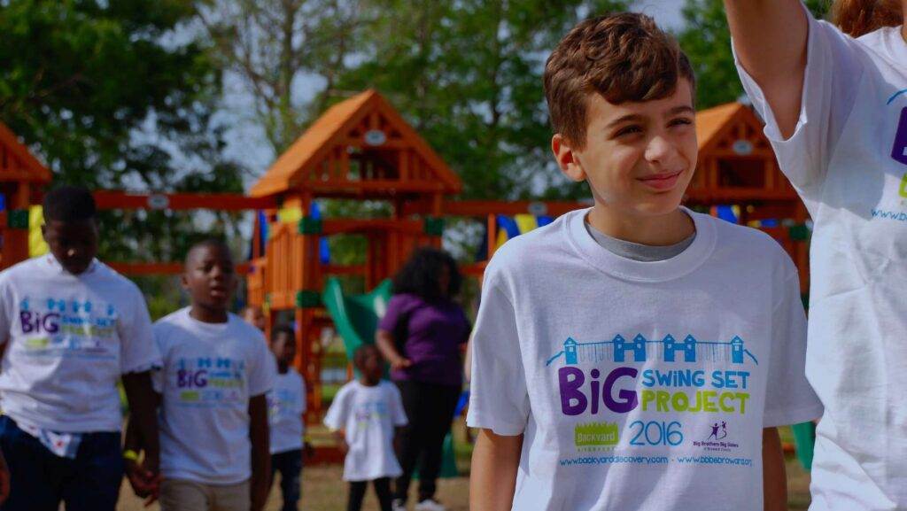 A group of children and adults wearing white "Big Swing Set Project 2016" T-shirts stand together in front of playground equipment on a sunny day, celebrating the installation of the longest swing set.