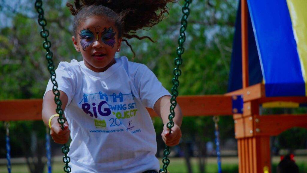 A child with a painted face and a white "Big Swing Set Project" t-shirt joyfully swings on the playground's longest swing set outdoors.