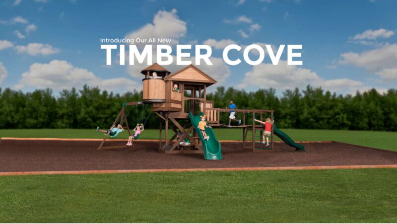 A wooden playset titled "Timber Cove" under a blue sky, with children engaged in various activities on it: swinging, sliding, and climbing. Boasting the longest swing set in the park, the playset is situated in an open grassy area with trees in the background.