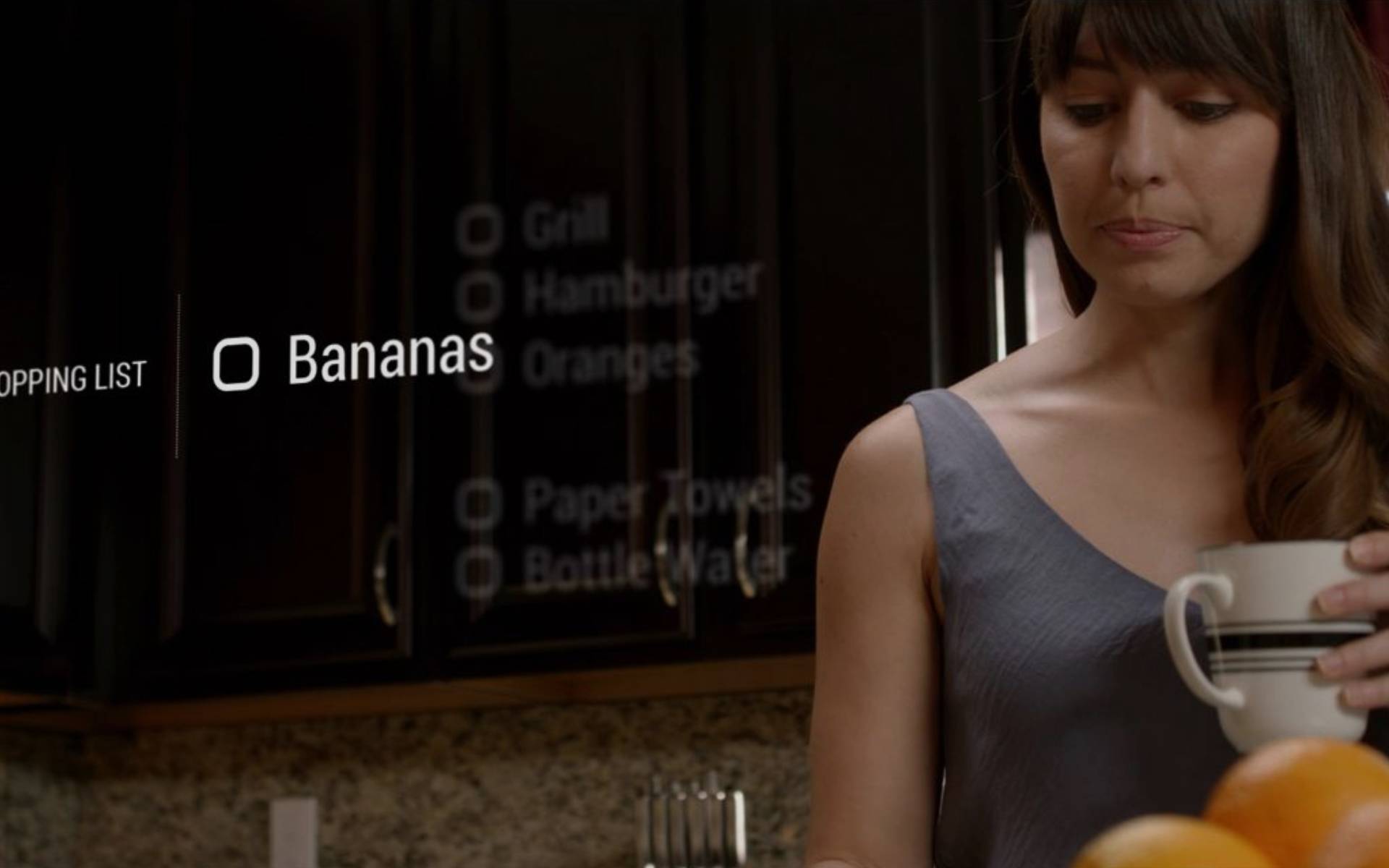 Woman in a kitchen holding a mug, standing next to a counter. A shopping list is visually projected, with "Bananas" checked off among other items.