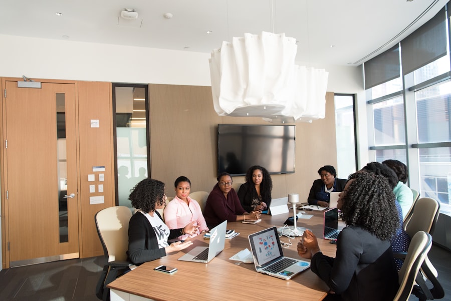 A group of people are seated around a conference table in a modern office, engaging in a meeting. Some have laptops open, and a large screen is mounted on the wall in the background.
