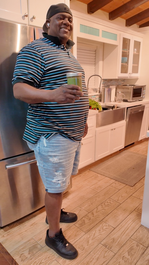 A person in a striped shirt and denim shorts stands in a kitchen holding a green morning smoothie, smiling. There are kitchen appliances and a bowl of produce in the background.