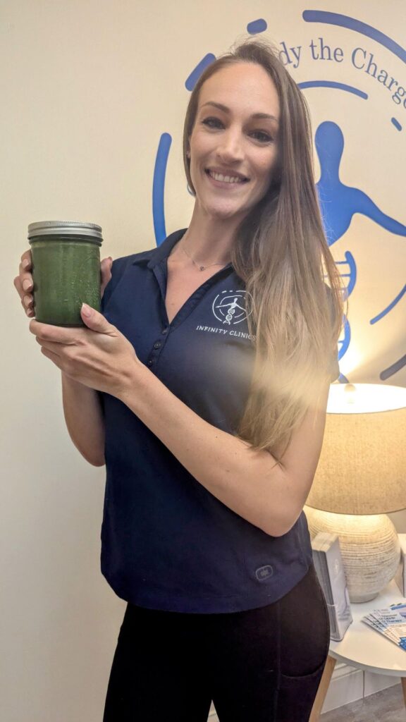A person in a navy shirt is holding a jar filled with a vibrant morning smoothie, standing proudly in front of a logo on the wall.