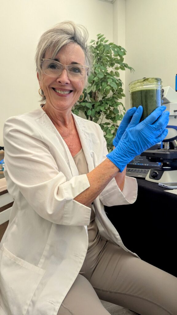 Woman in lab coat and gloves holding a jar with green liquid, seated near a microscope, smiling at the camera as if she's just discovered the secret to a perfect morning smoothie.