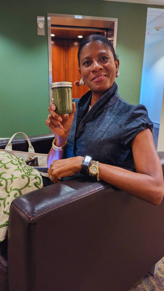 Woman seated on a brown couch holds up a jar filled with her morning smoothie, smiling at the camera. She wears a dark top, a watch, and sits beside a patterned pillow and a beige bag.