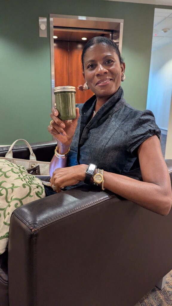A woman sits on a couch holding up a jar filled with a green morning smoothie, looking at the camera and smiling.