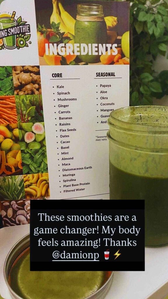 A glass of green smoothie sits on a table next to a small bowl and a printed ingredients list featuring various fruits and vegetables.