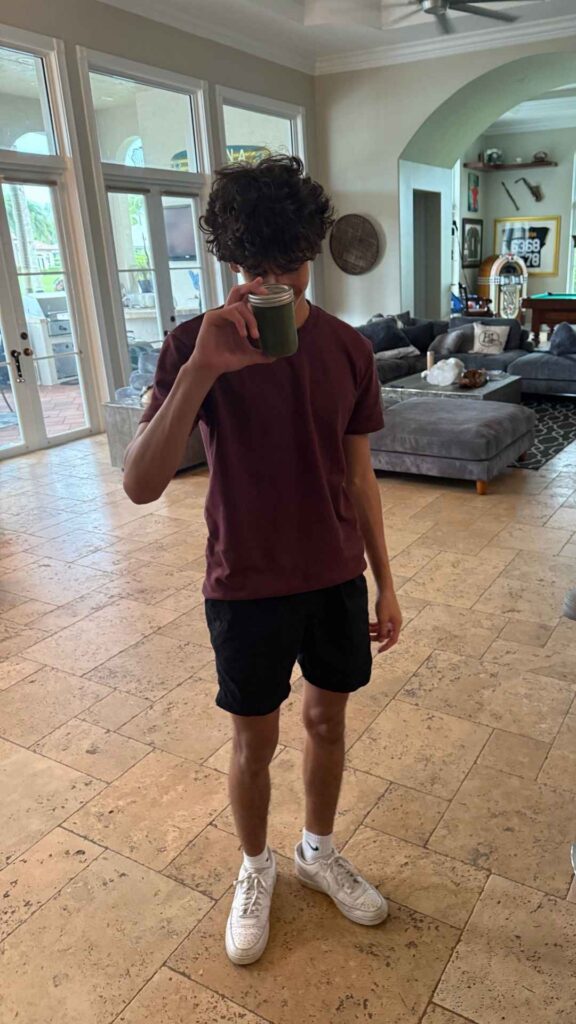 A young person in a maroon shirt, black shorts, and white sneakers stands indoors, holding a glass with a green morning smoothie in front of their face.