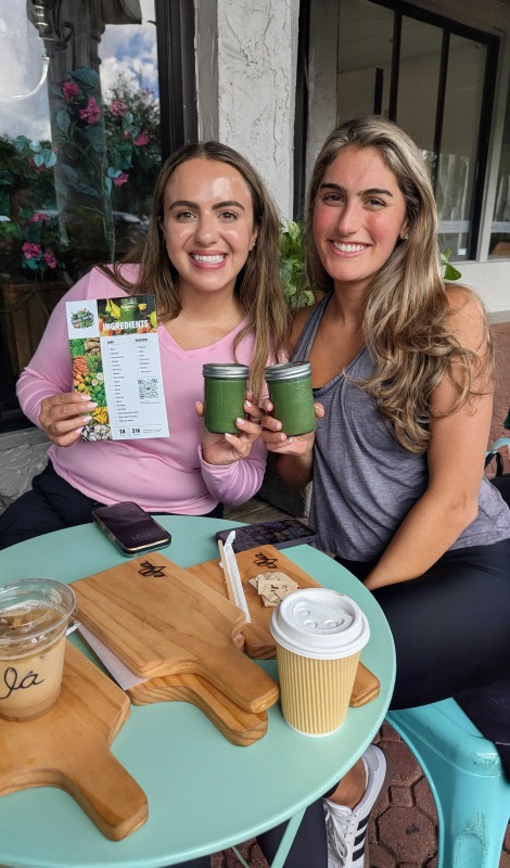 Two women sit at an outdoor table, smiling and holding morning smoothies, with a menu and coffee cups on wooden boards in front of them.