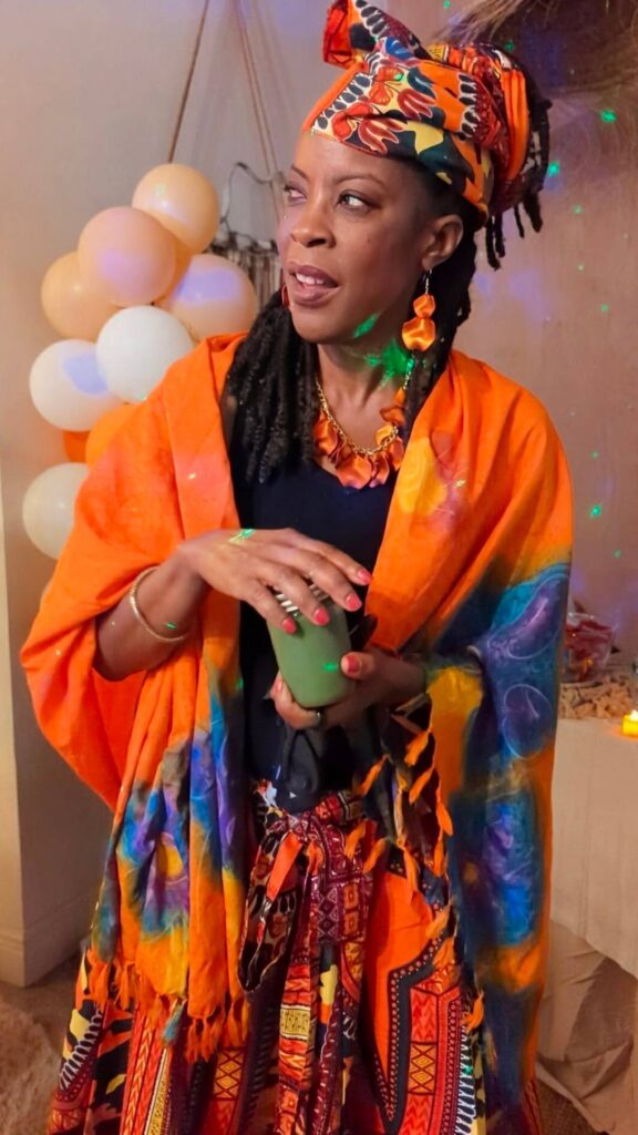 A woman wearing a colorful, patterned orange outfit and headwrap holds a green morning smoothie, standing indoors with balloons and party decorations in the background.
