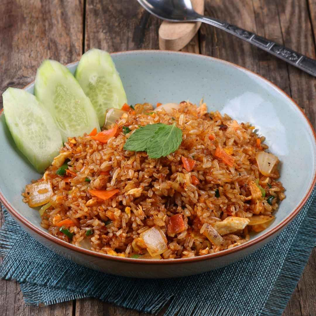 A bowl of fried rice with diced vegetables, garnished with a mint leaf and cucumber slices, sits on a blue cloth with a metal spoon nearby—perfect for those craving delicious easy eats.