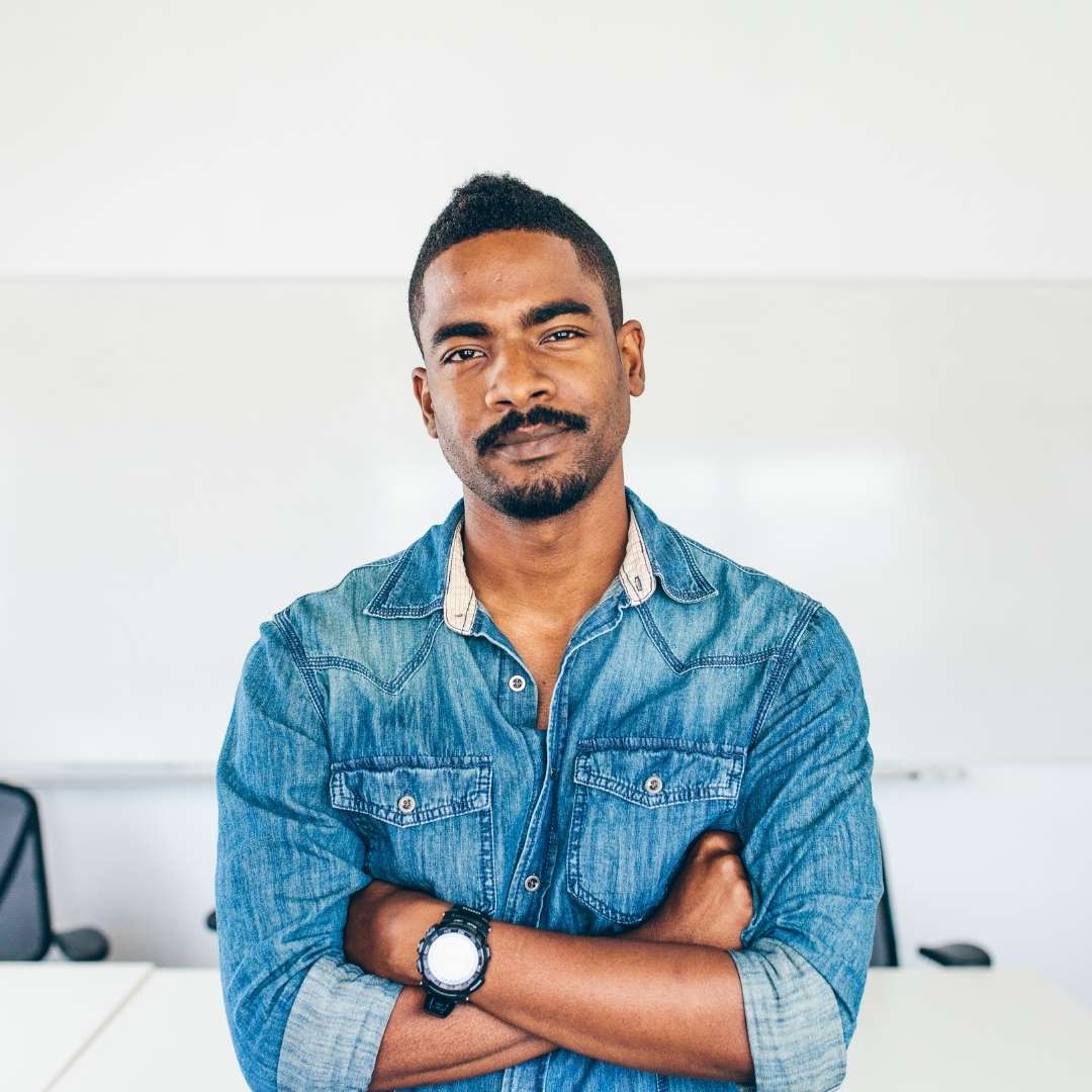A man standing indoors with arms crossed, wearing a denim shirt and watch, in front of a whiteboard and office chairs, brainstorming ideas for easy eats.