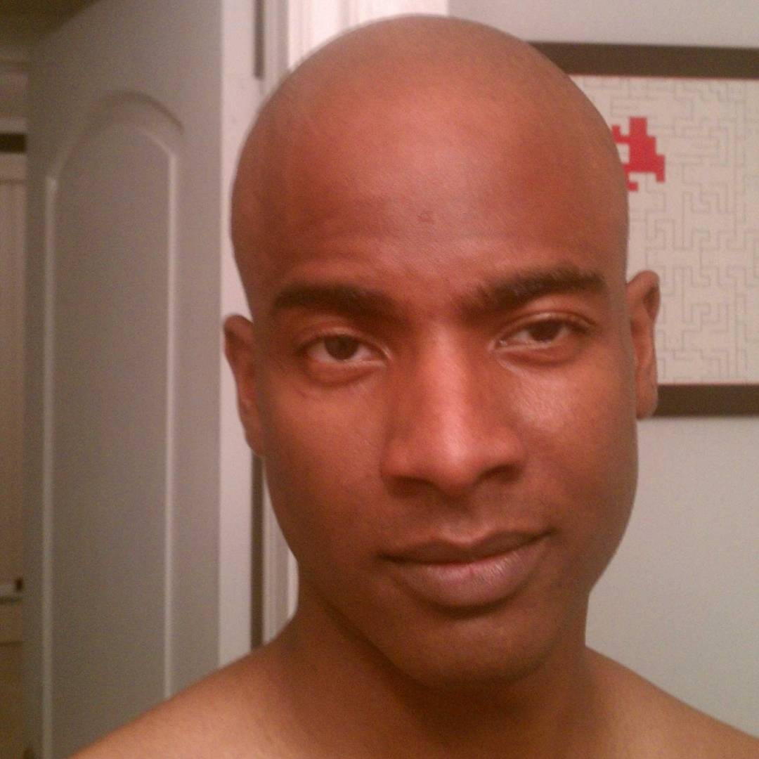 A person with a shaved head is standing indoors, looking at the camera. In the background, a partially open door, framed artwork, and a hint of easy eats create a relaxed atmosphere.