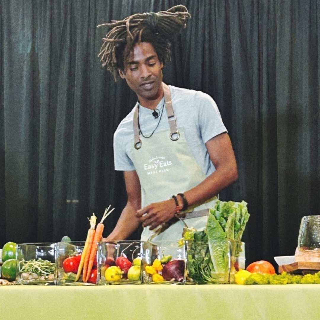 A man wearing an apron stands behind a table with various fresh vegetables and greens on display, preparing easy eats in front of a black curtain backdrop.